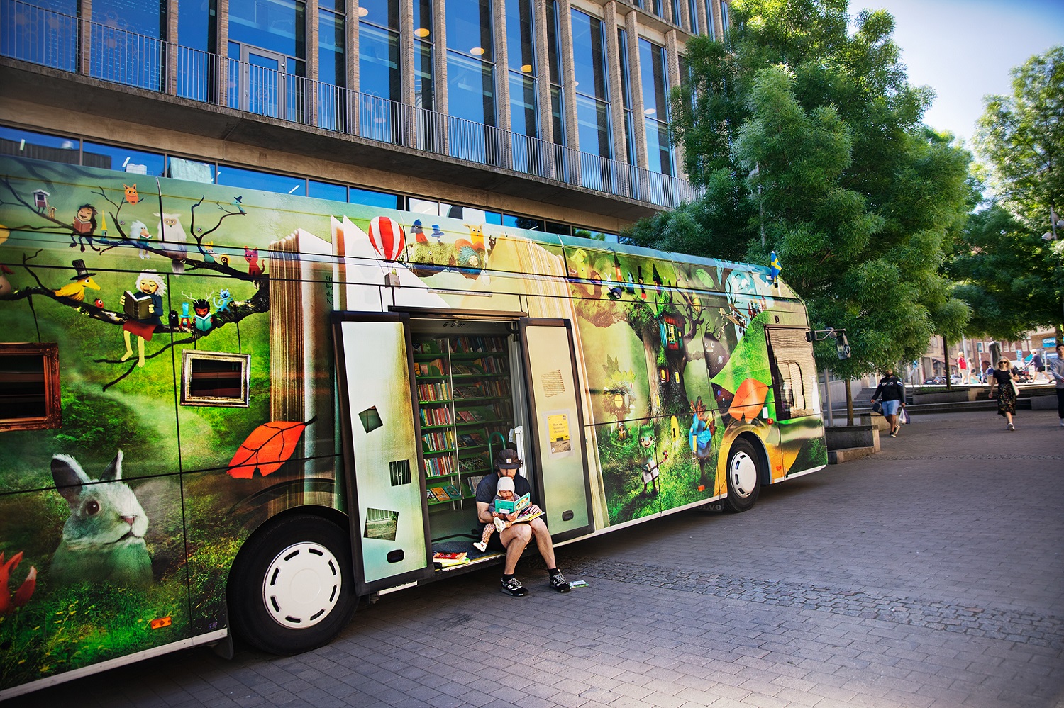 A casually dressed man holds a young infant in the doorway of a brightly painted bus on a pedestrian street.