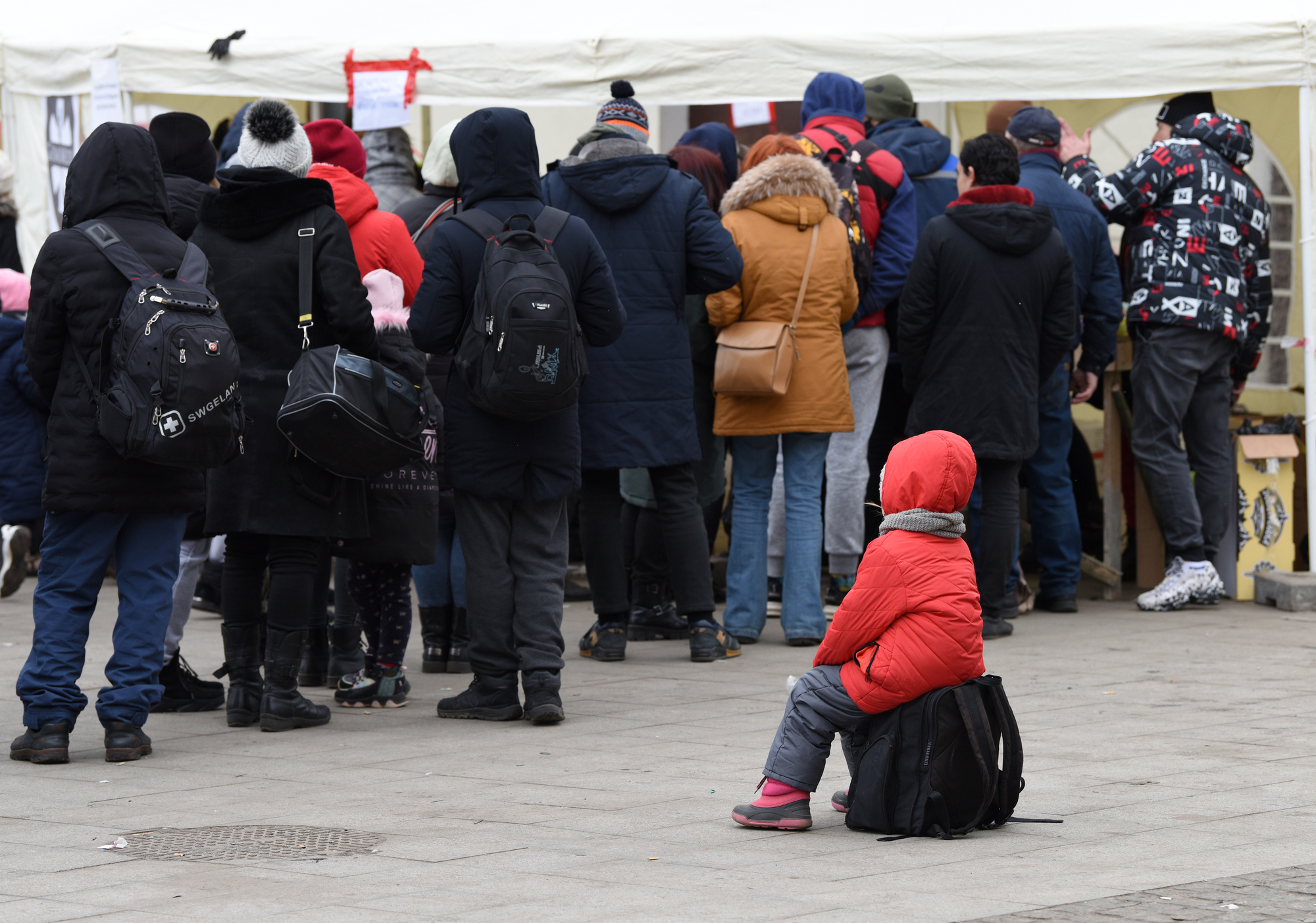 Child in red jacket sits on suitcase near line of evacuees in Lviv, Ukraine