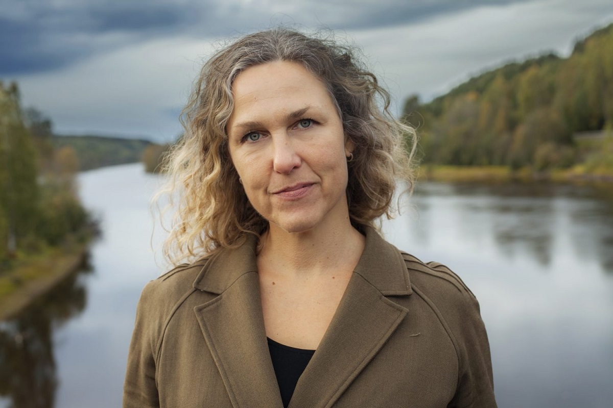 Marit Kapla wearing an olive blazer standing in front of a lake setting.