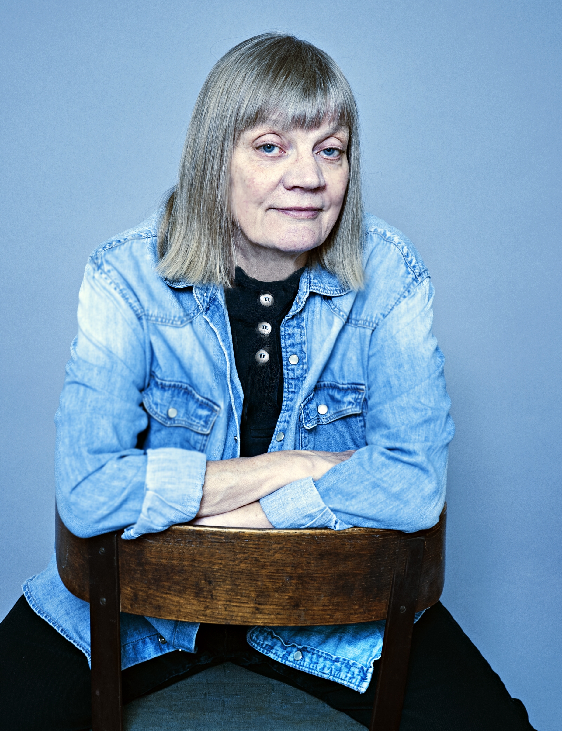 Gun-Britt Sundström wearing a denim jacket with a short blond bob, sitting backwards on a chair