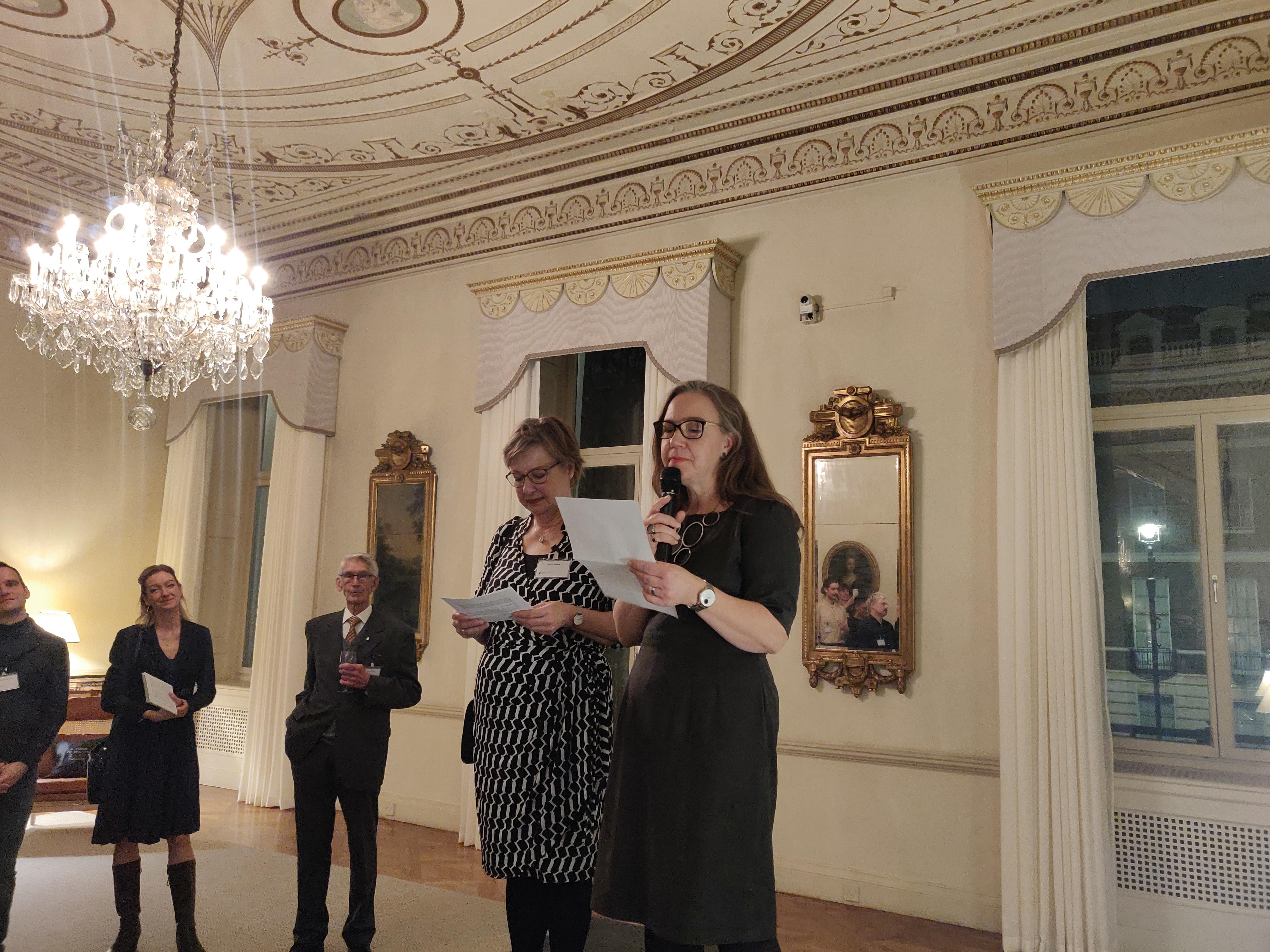 Two women read a speech on a stage while audience members look on