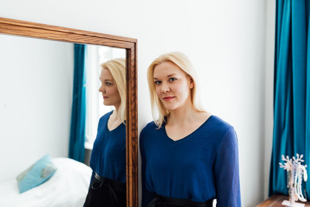 Blond haired woman in a blue top standing sideways to a mirror.