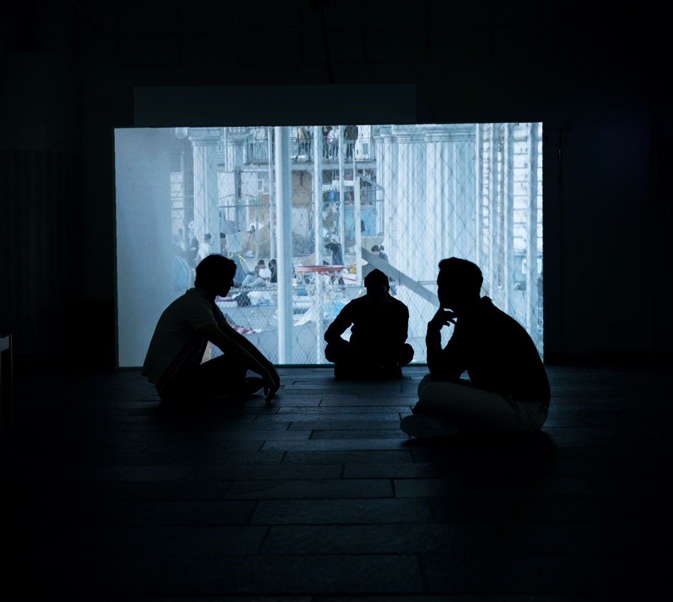 Three performers sit in darkness as projection is cast across back wall.