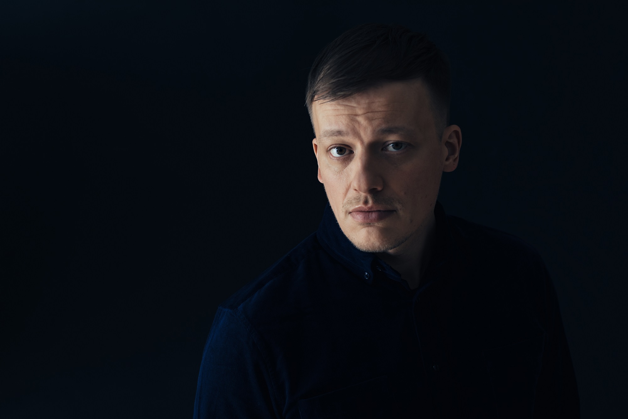 Photo of young man with dark shirt and hair stepping out of the shadows.