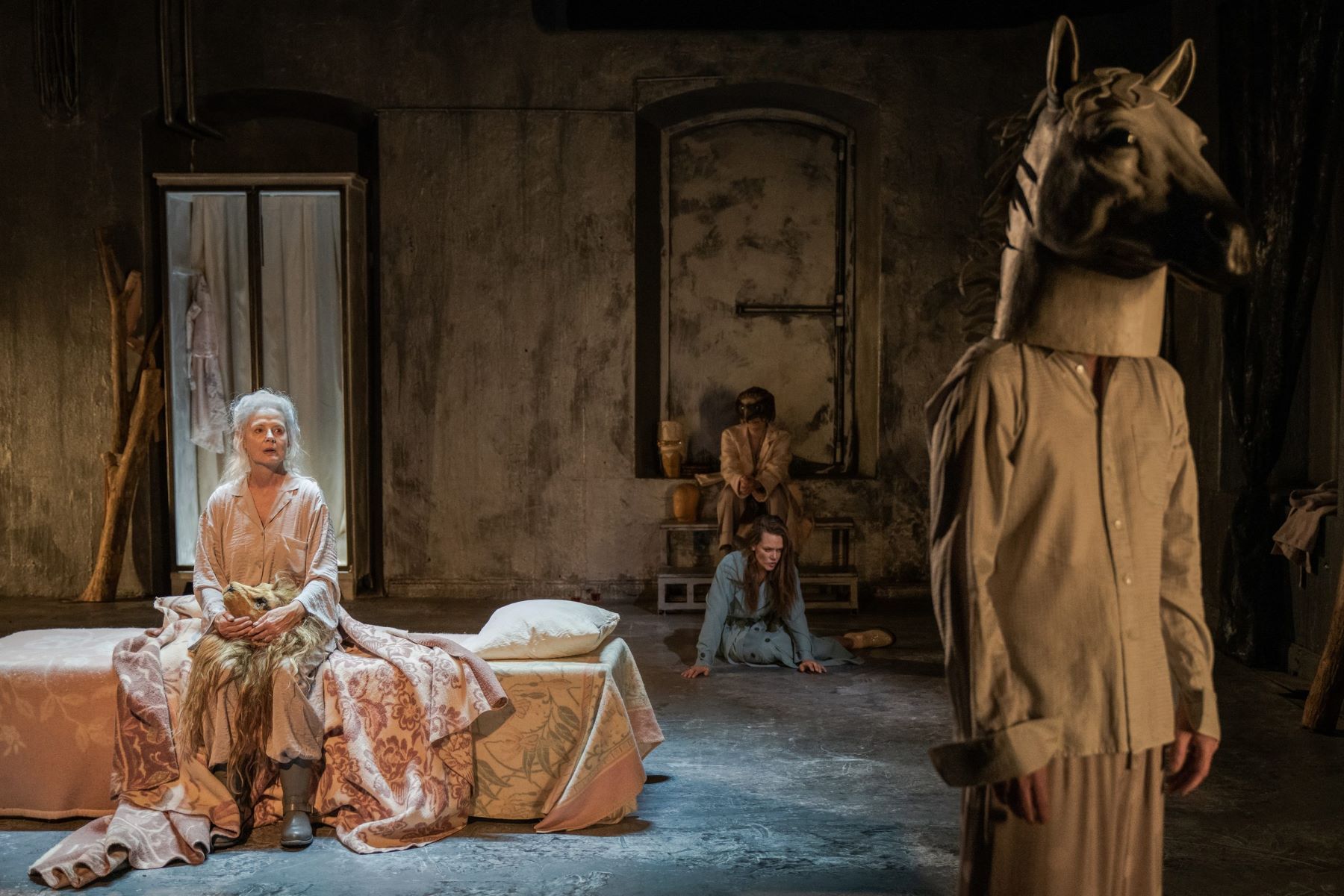 An actor sits on a bed, staring at another performer wearing a horse's head.