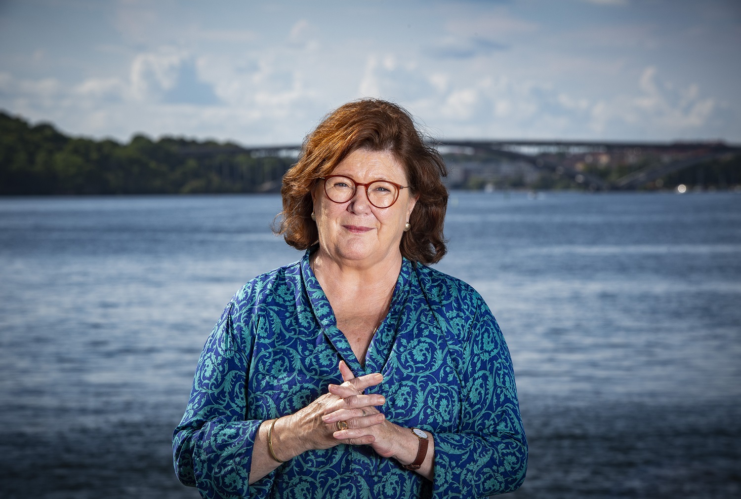 Woman with shoulder-length brown hair and glasses squints into camera in front of urban lake landscape.
