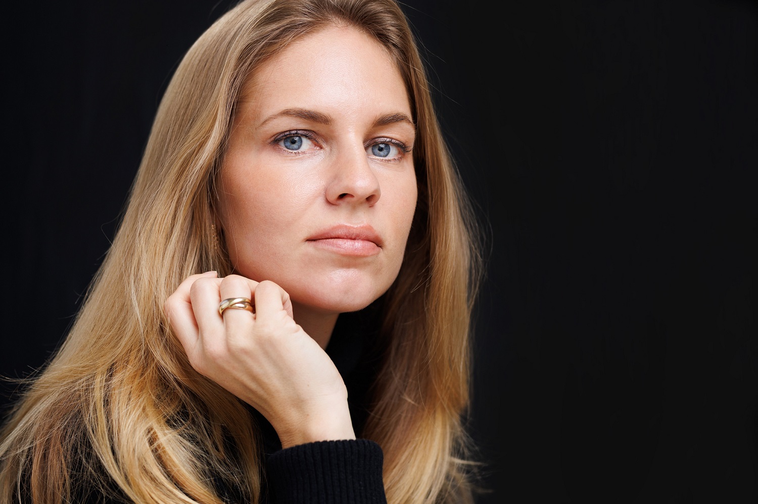 Young woman with blonde hair and blue eyes wearing a black jumper looks into camera.