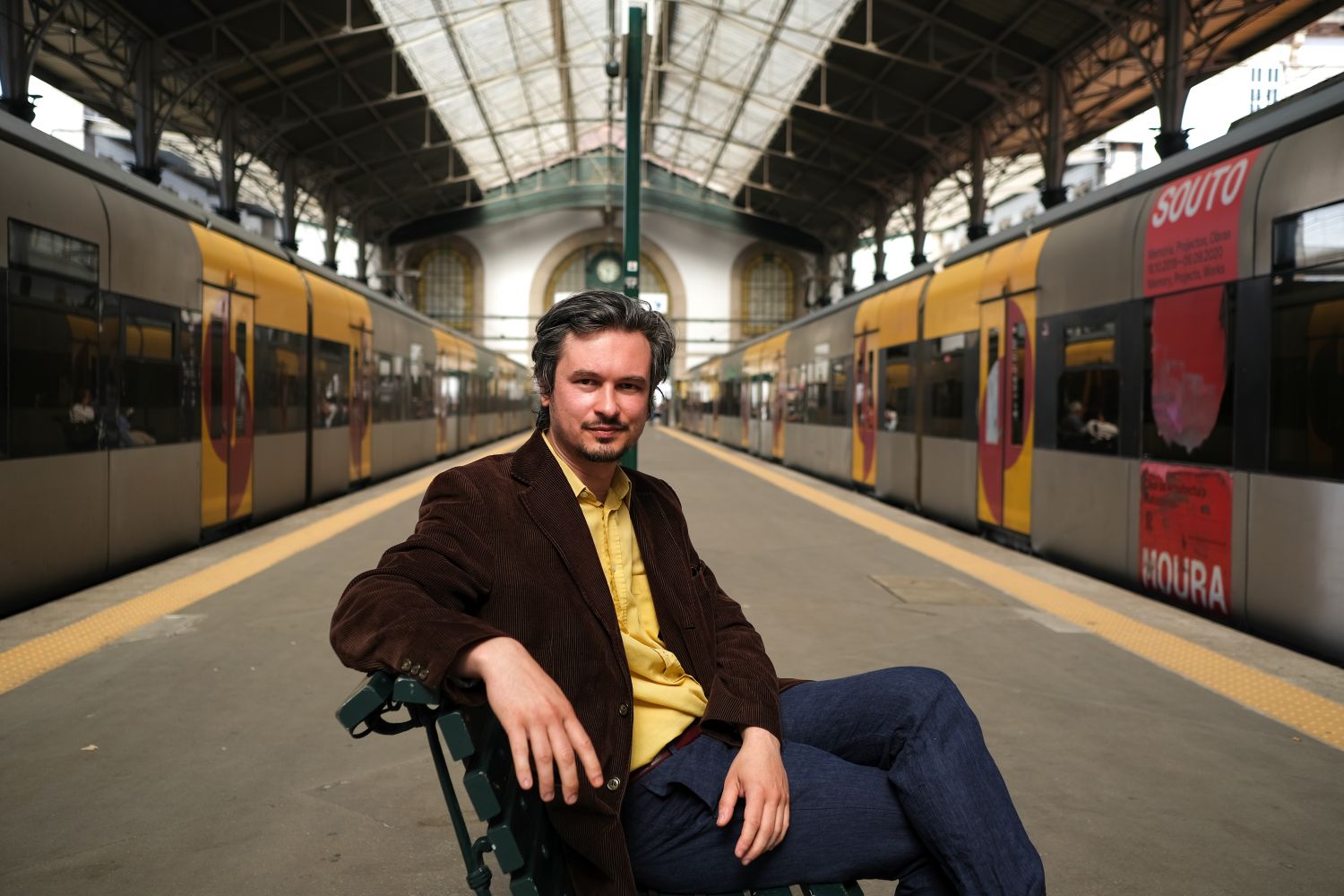 Maxim Grigoriev sitting on bench on train platform flanked by trains