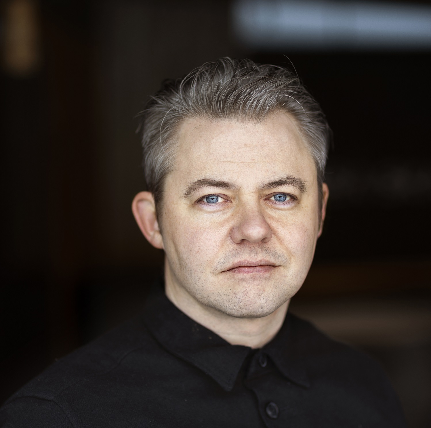 Photo of grey-haired man with pale blue eyes in black shirt against a blurred background