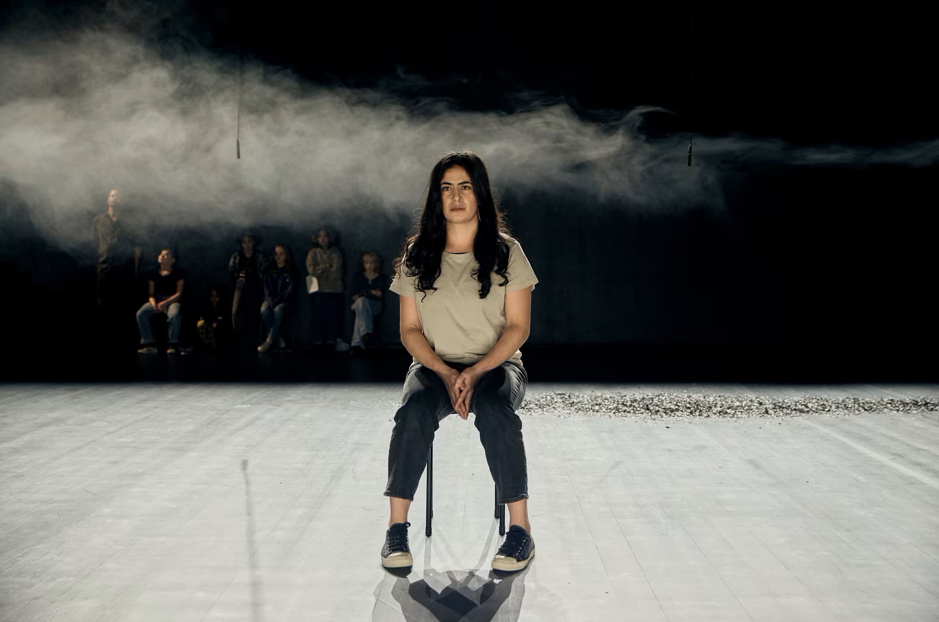 An actor sits on a chair on a darkened stage, with a chorus behind her.