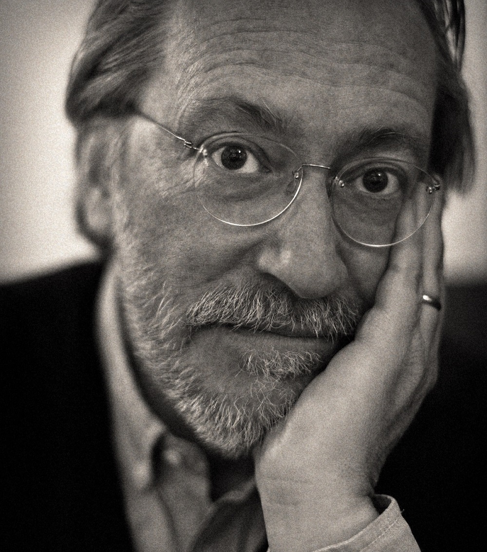 Black-and-white photo of grey-haired man in glasses resting his head in his hand.