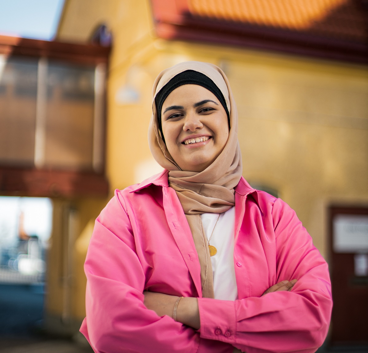 Young woman wearing peach-coloured hijab and a pink jacket smiles at the camera in front of yellow building.