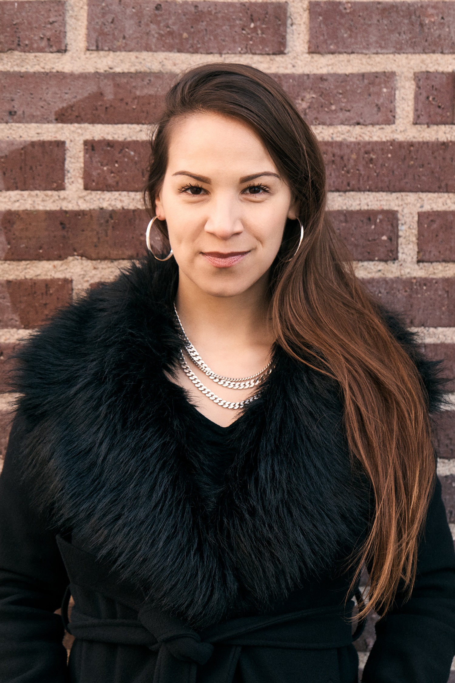 Young woman with long dark hair wearing a fluffy dark coat smiles at the camera with a brickwork background