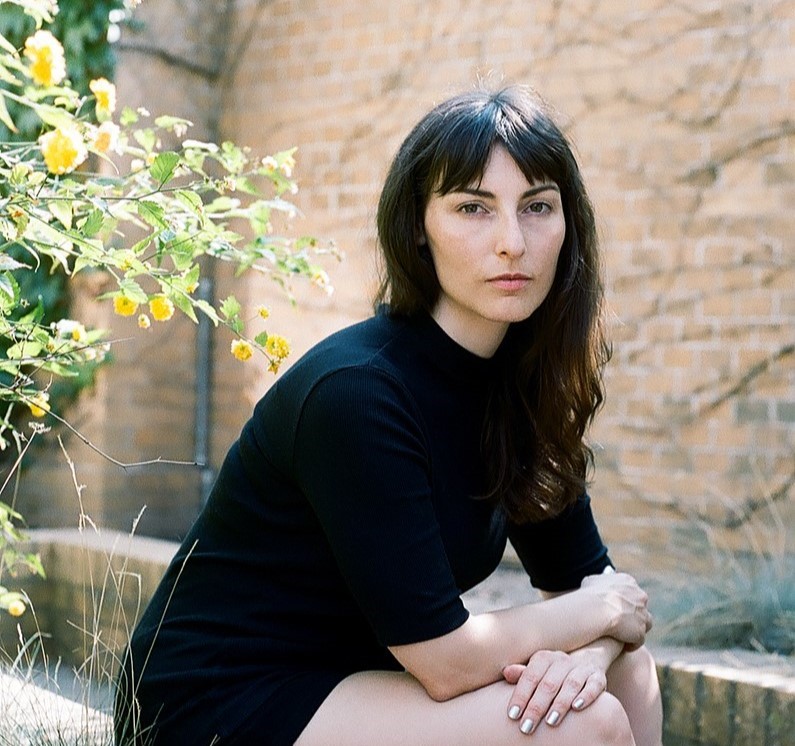 Saskia Vogel in black dress sitting on brick wall in walled garden.