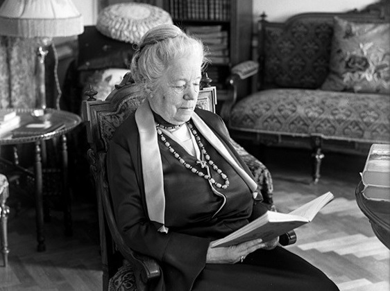 Black-and-white photo of elderly woman sitting in a chair reading.