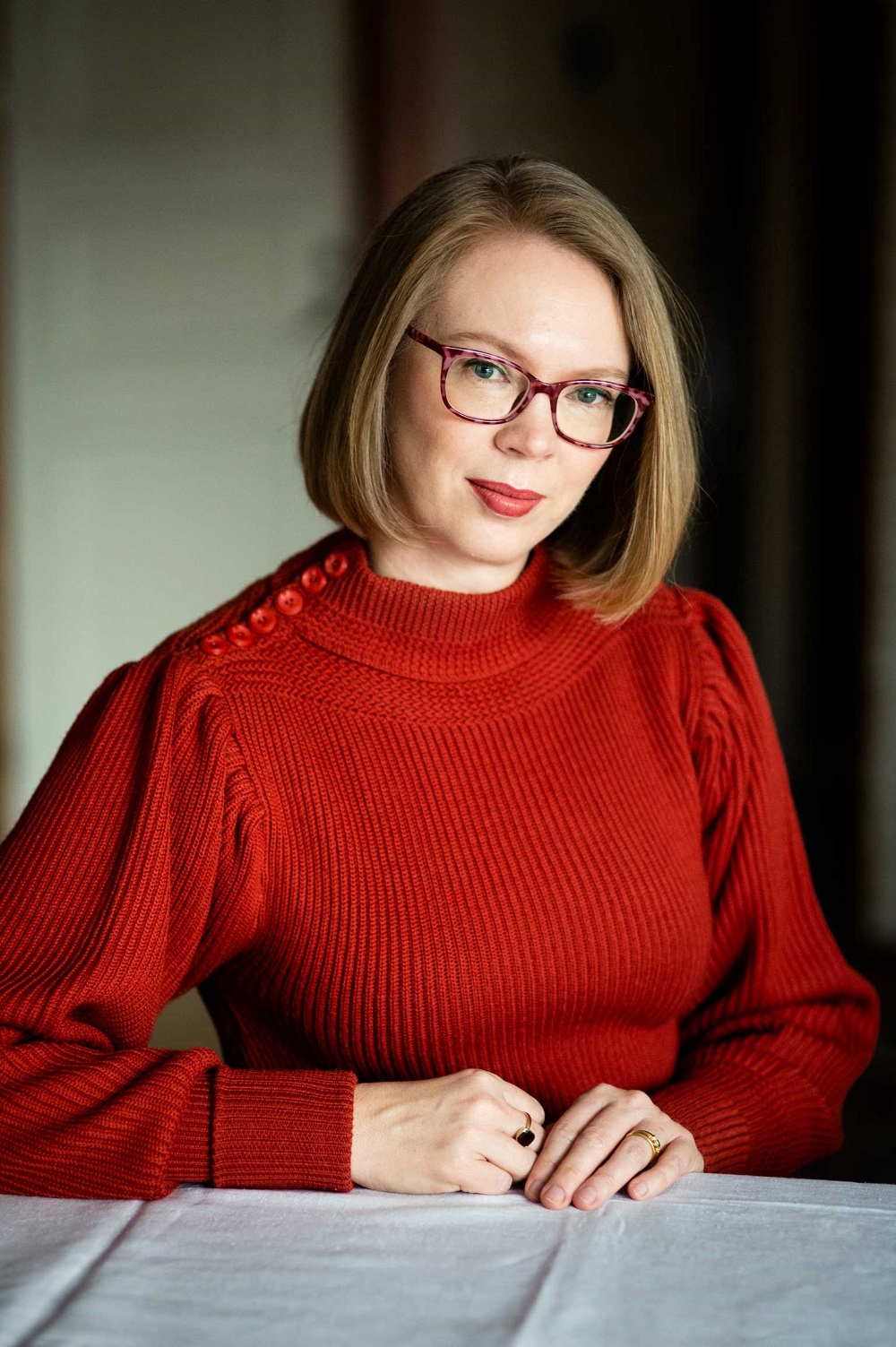 Young woman with reddish-blonde hair in a bob wearing glasses and a red jumper looks into camera.