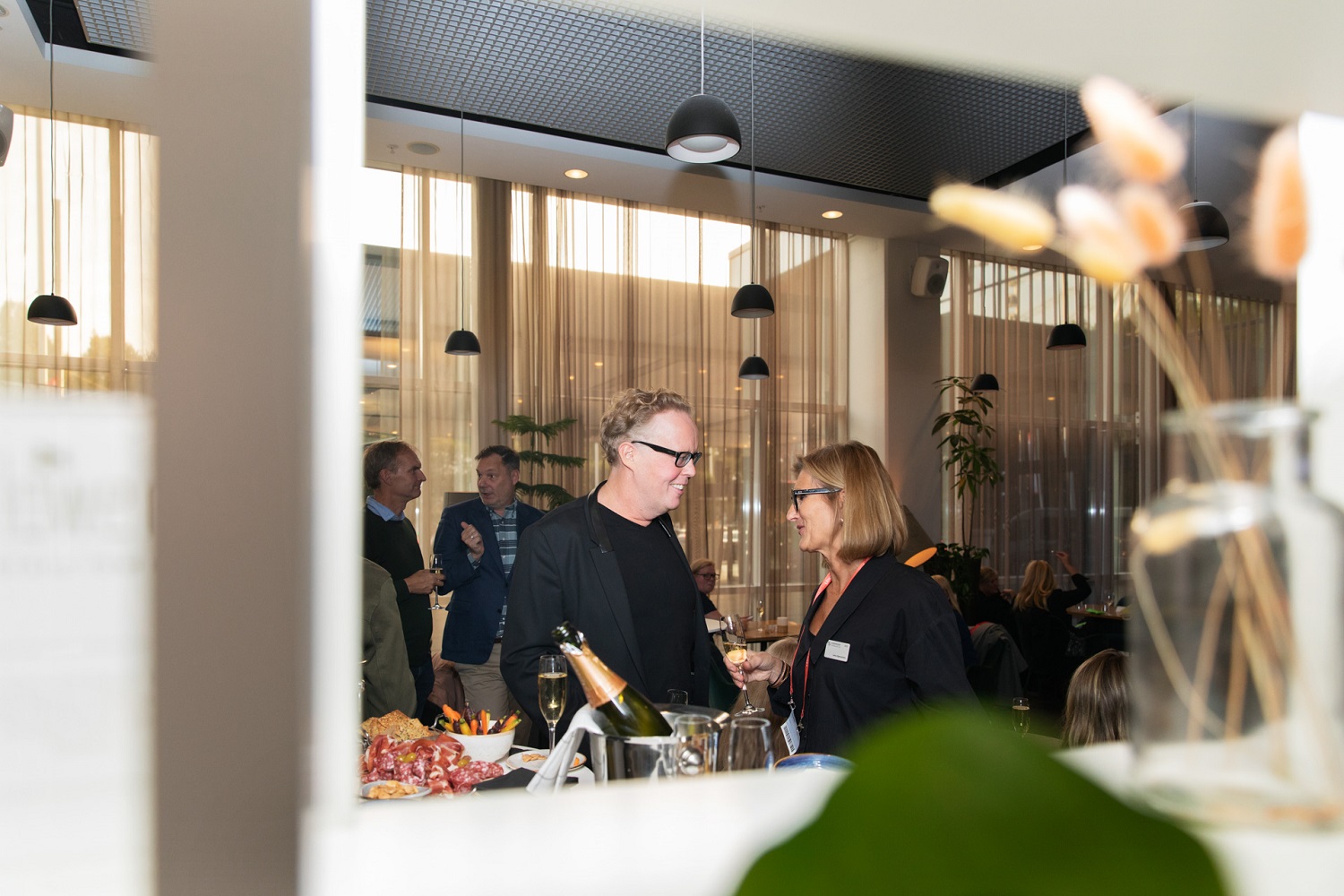 Tall man in a black suit and blond woman in black dress chat over canapes.