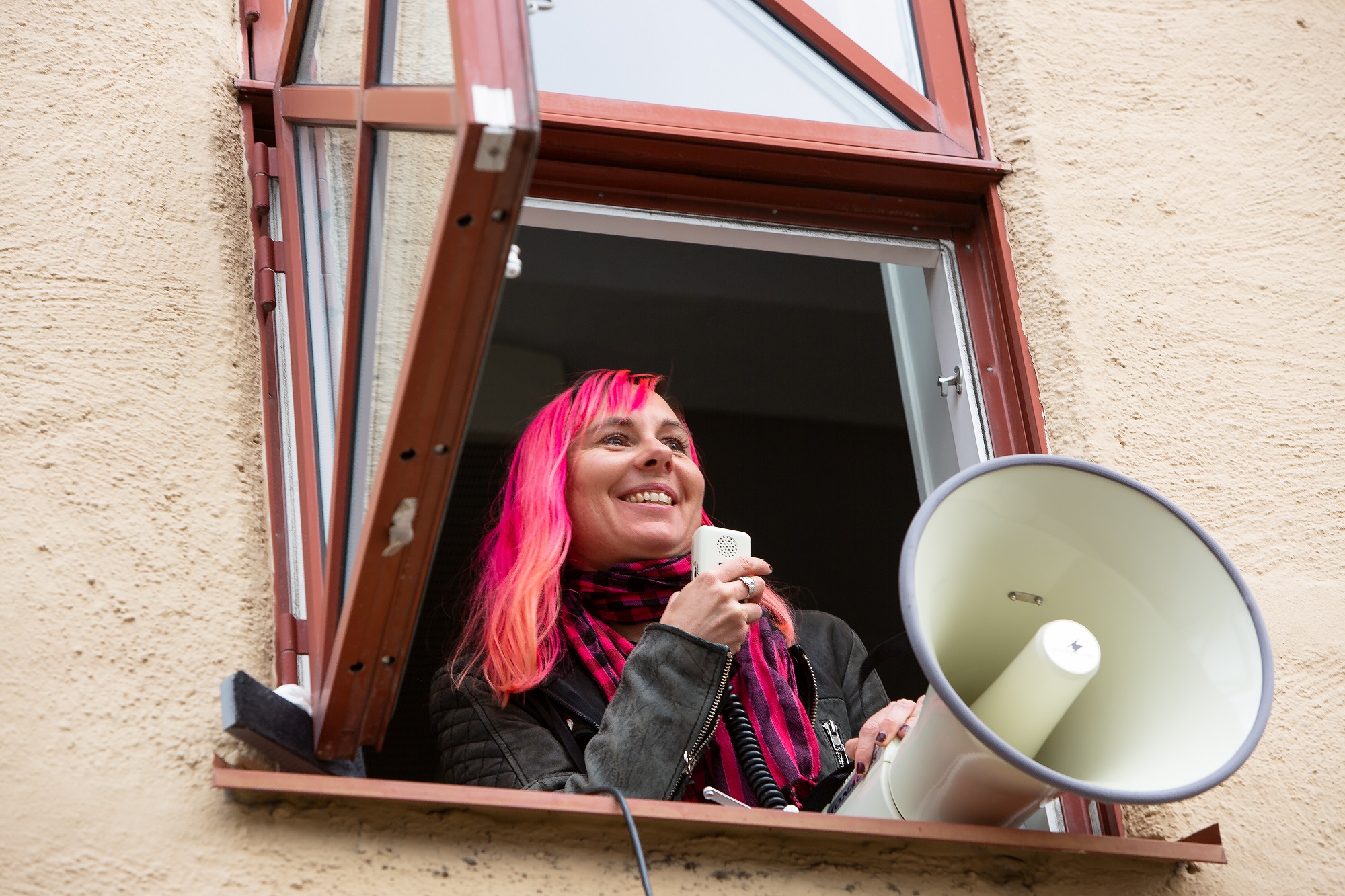 A smiling woman with pink hair holds a megaphone through an open window.