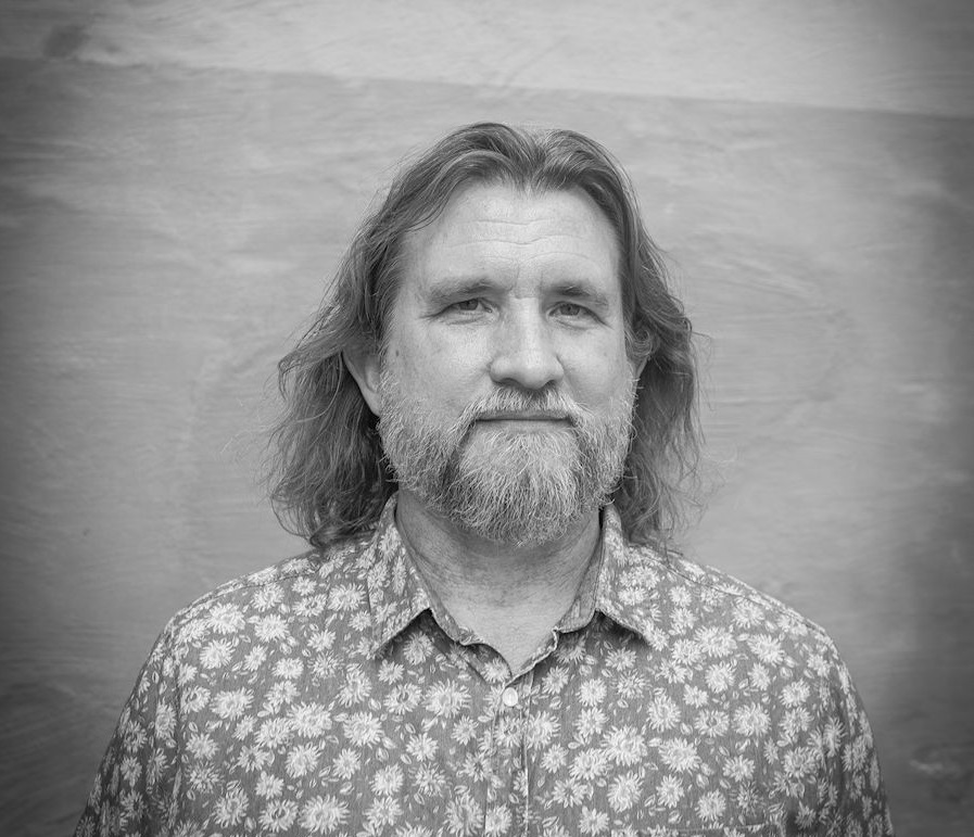 Black-and-white photo of a man with long hair and a beard, wearing a flower-print shirt.