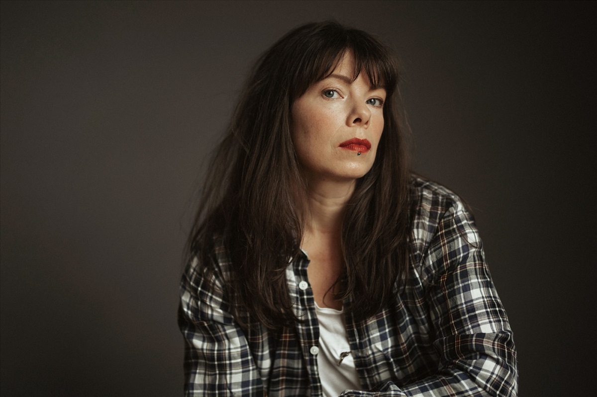 Woman with long brown hair in chequered shirt looking askance at camera.