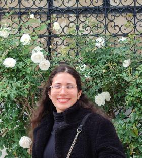 Hakima Choukri wearing black clothes and glasses standing in front of a flower bush.