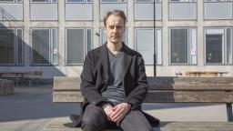 Man in grey clothing sitting on wooden bench in front of building