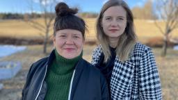Two women in coats looking into camera in front of an open field.