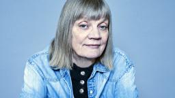 Gun-Britt Sundström wearing a denim jacket with a short blond bob, sitting backwards on a chair
