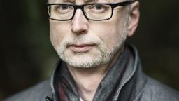 Grey haired man in glasses wearing jacket and scarf.