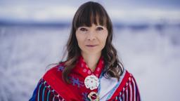 Woman with dark hair in traditional Sami dress standing in front of blue-tinged snowy background.