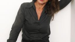 Woman with long dark hair and green earrings stands with her hand on her head against white background.