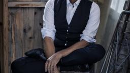 Man with grey hair and blue eyes in shirt and waistcoat sits in a wooden cottage with a mirror in the background.