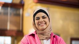 Nora Khalil wearing pale hijab and pink blazer smiles in front of yellow building.