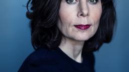 Woman with green eyes and dark hair wearing plum lipstick in front of a blue background.