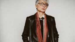 Woman with short grey hair, glasses and leather jacket standing against a white background.