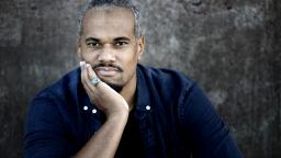 Johannes Anyuru in a denim shirt sits with head resting on his hand, wearing green ring.