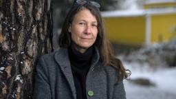 Lotta Geffenblad stands next to tree trunk wearing grey coat and glasses.