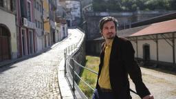 Maxim Grigoriev in shirt and blazer leaning against a metal fence on a cobbled street.