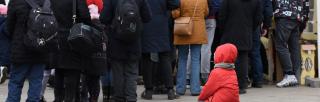 Red child sits on suitcase near queue of evacuees in Lviv.