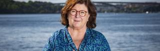 Woman with shoulder-length brown hair and glasses squints into camera in front of urban lake landscape.