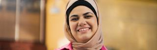 Young woman wearing peach-coloured hijab and a pink jacket smiles at the camera in front of yellow building.