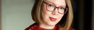 Young woman with reddish-blonde hair in a bob wearing glasses and a red jumper looks into camera.